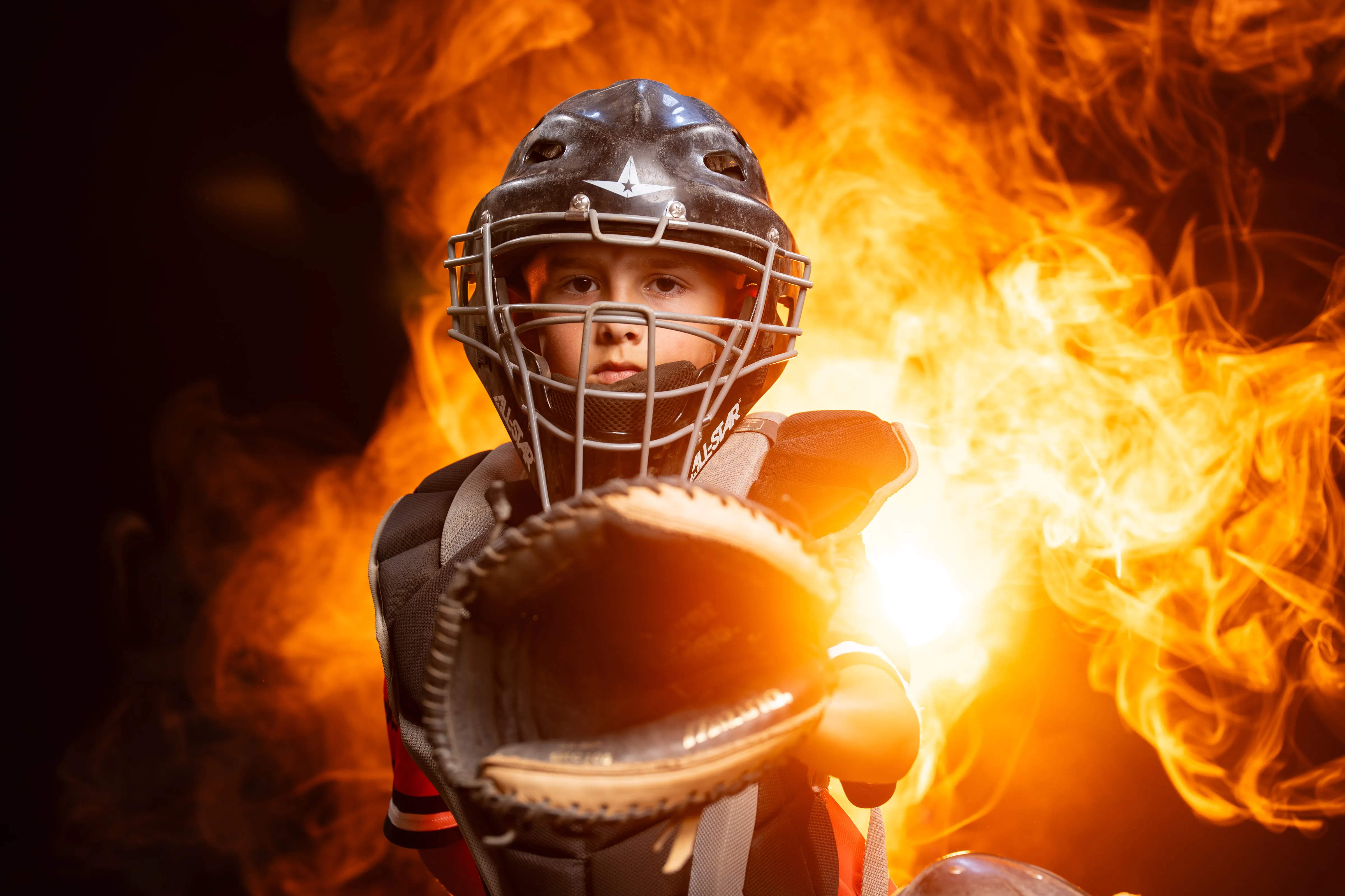 Youth baseball catcher in full gear holding a glowing ball of fire, dramatic studio sports portrait