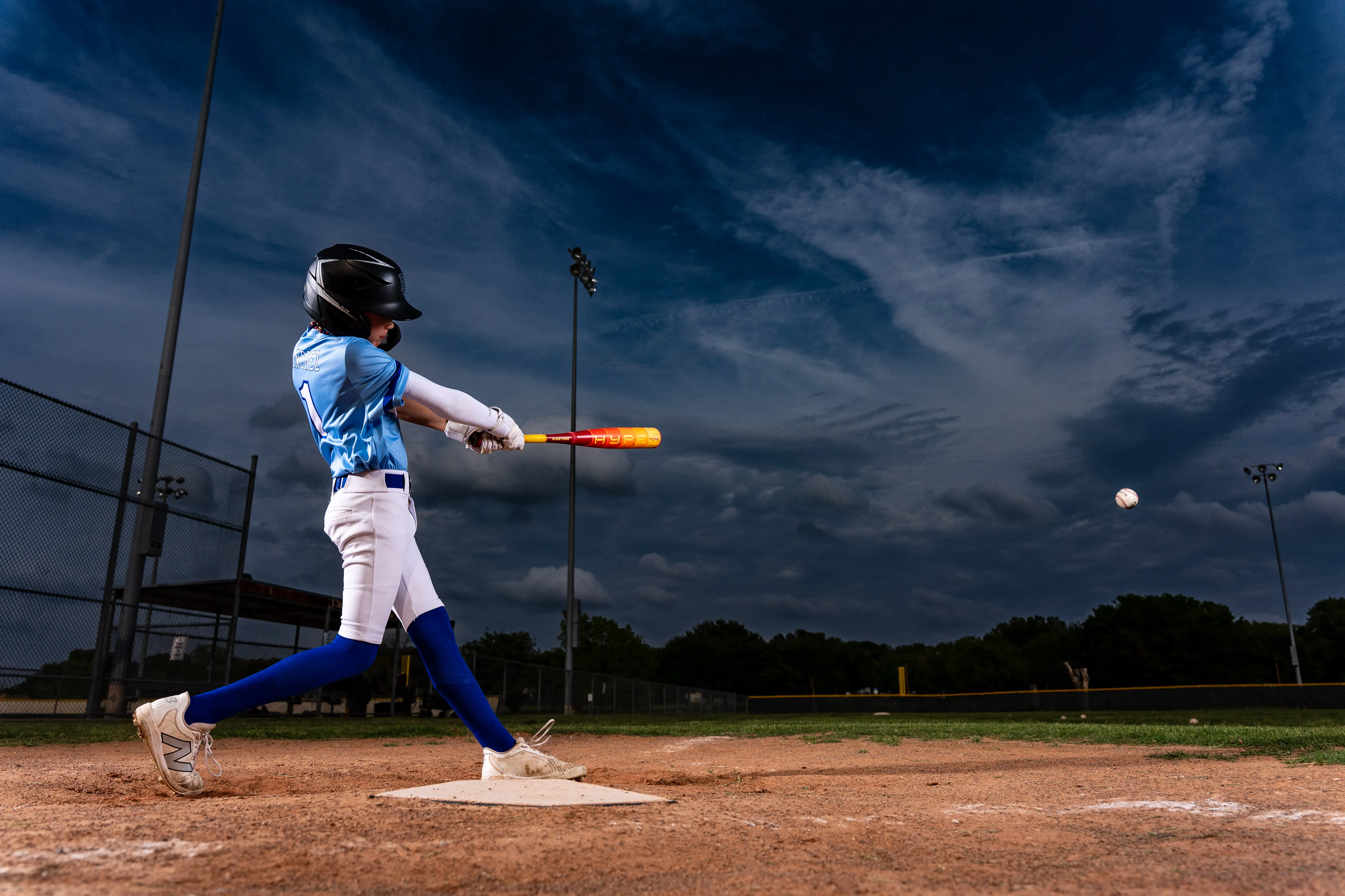 Baseball batter mid-swing at dusk under a dramatic sky on the field